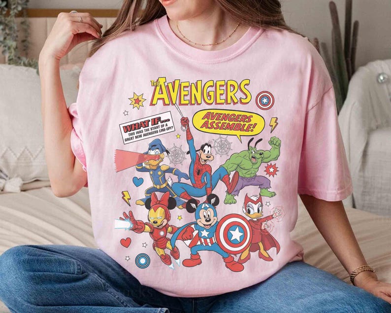 May include: A pale pink t-shirt with a comic book style design of Disney characters as Avengers. The graphic includes the words "AVENGERS" and "AVENGERS ASSEMBLE!" and other text. The shirt has a round neck and short sleeves.