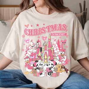 May include: Cream-colored t-shirt featuring a pink graphic with the text "CHRISTMAS ON MAIN STREET." The design includes cartoon characters, a castle, Christmas trees, and gift boxes, creating a festive holiday theme.