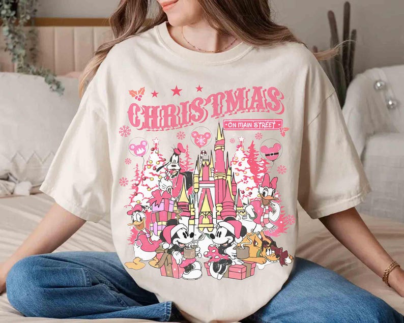 May include: Cream-colored t-shirt featuring a pink graphic with the text "CHRISTMAS ON MAIN STREET." The design includes cartoon characters, a castle, Christmas trees, and gift boxes, creating a festive holiday theme.