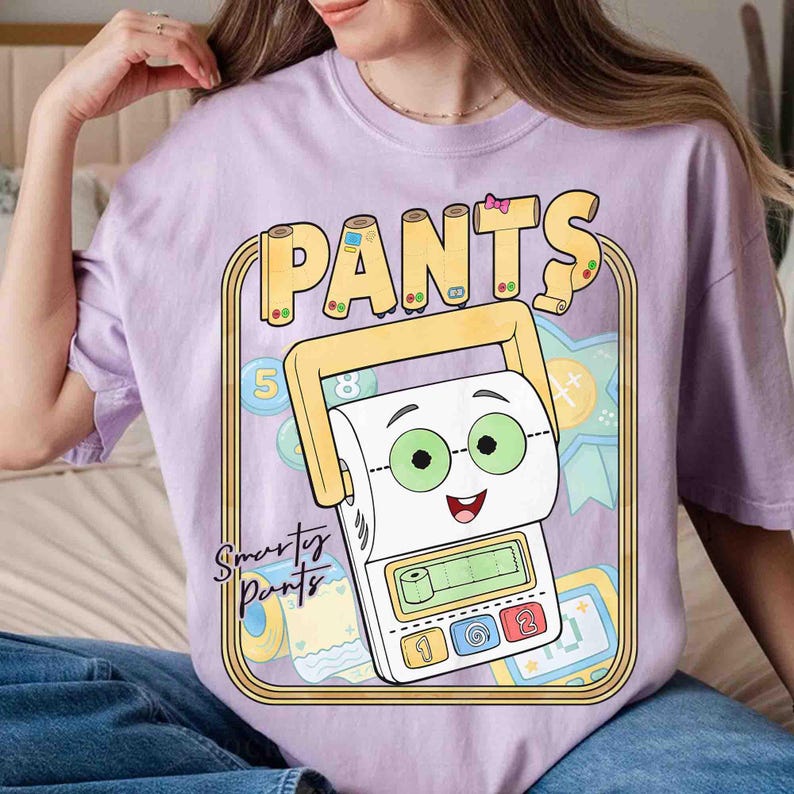 May include: A light purple t-shirt featuring a graphic of a cartoon toilet paper roll character wearing green glasses. The design includes the text "PANTS" at the top and "Smarty Pants" on the side, surrounded by a yellow border and school-themed icons.