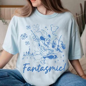 Puede incluir: Camiseta azul claro con una ilustraci&oacute;n azul de Mickey Mouse con un sombrero de mago, sosteniendo una varita y cubos, con un castillo y fuegos artificiales. La palabra "Fantasmic!" est&aacute; escrita en cursiva debajo.