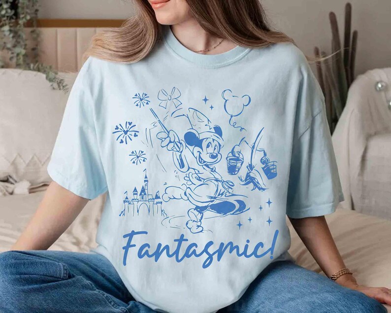 Puede incluir: Camiseta azul claro con una ilustraci&oacute;n azul de Mickey Mouse con un sombrero de mago, sosteniendo una varita y cubos, con un castillo y fuegos artificiales. La palabra "Fantasmic!" est&aacute; escrita en cursiva debajo.