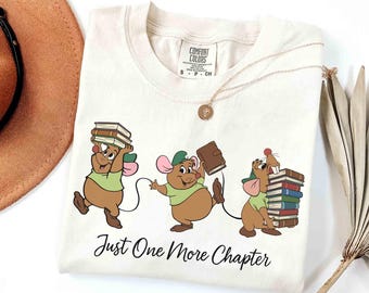 May include: Cream-colored t-shirt featuring a cartoon illustration of three mice carrying books. The mice are wearing green shirts and hats. The text "Just One More Chapter" is printed below the illustration. A gold necklace is draped across the shirt.