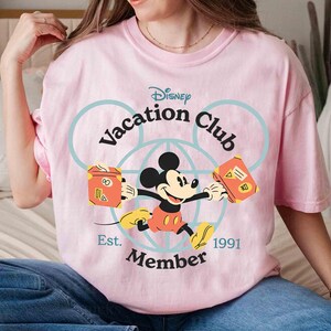 Puede incluir: Camiseta rosa claro con Mickey Mouse del Disney Vacation Club. Mickey sostiene dos maletas naranjas. El texto "Disney Vacation Club Member Est. 1991" est&aacute; en azul en la camiseta.