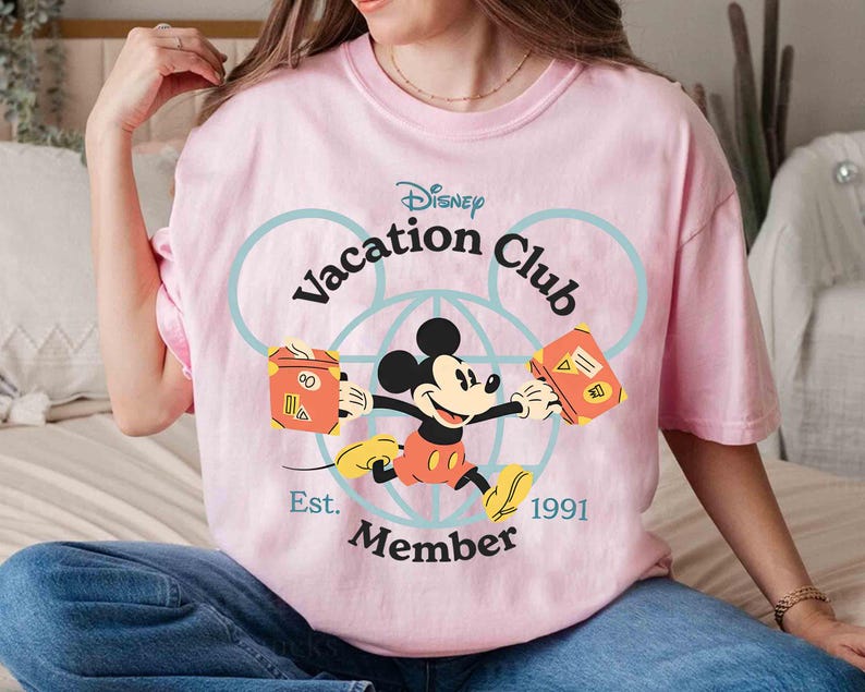 Puede incluir: Camiseta rosa claro con Mickey Mouse del Disney Vacation Club. Mickey sostiene dos maletas naranjas. El texto "Disney Vacation Club Member Est. 1991" est&aacute; en azul en la camiseta.