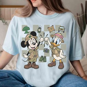 May include: Light blue t-shirt featuring Mickey Mouse and Daisy Duck in safari outfits, giving a high five. The design includes safari-themed elements like a jeep, binoculars, and a camera. The background is a checkered pattern.
