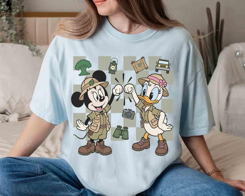 May include: Light blue t-shirt featuring Mickey Mouse and Daisy Duck in safari outfits, giving a high five. The design includes safari-themed elements like a jeep, binoculars, and a camera. The background is a checkered pattern.