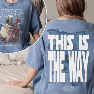 May include: A blue t-shirt featuring a graphic of The Mandalorian and Grogu on the front with the text "STAR WARS" and "THE MANDALORIAN AND GROGU". The back of the shirt displays the text "THIS IS THE WAY" in large white letters over a blue design.