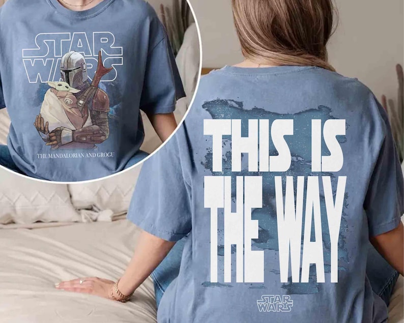 May include: A blue t-shirt featuring a graphic of The Mandalorian and Grogu on the front with the text "STAR WARS" and "THE MANDALORIAN AND GROGU". The back of the shirt displays the text "THIS IS THE WAY" in large white letters over a blue design.