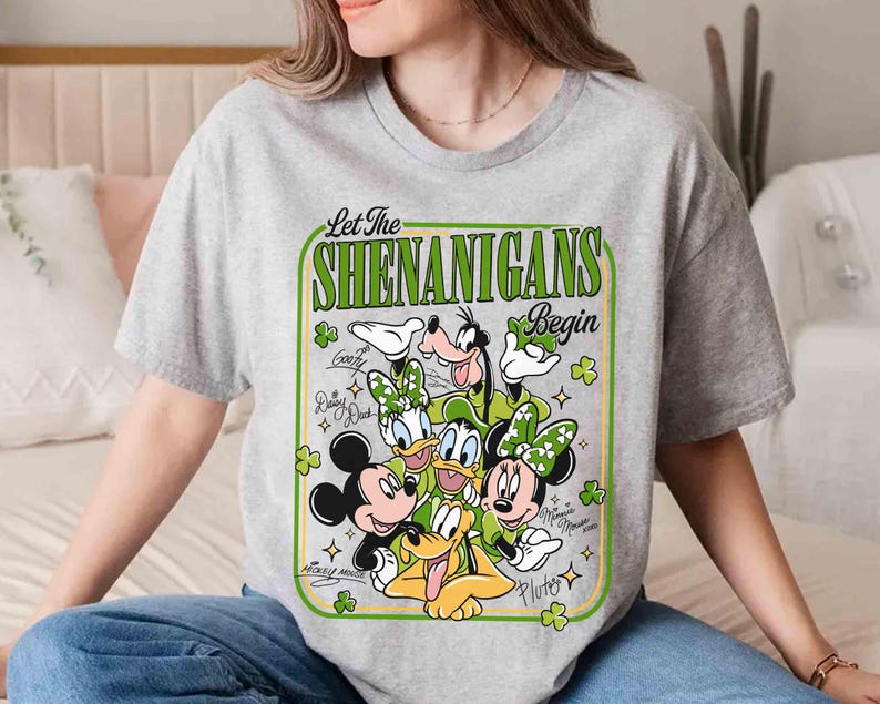 May include: A gray t-shirt featuring a St. Patrick's Day design with Mickey Mouse, Minnie Mouse, Donald Duck, Daisy Duck, Goofy, and Pluto. The text "Let The Shenanigans Begin" is in a green frame with shamrocks.