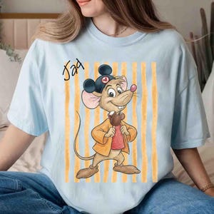 May include: A light blue t-shirt featuring a graphic of a cartoon mouse wearing a black hat with ears, a yellow jacket, and a red shirt, holding a brown treat. The text "Jad" is printed in black script above the mouse, set against yellow stripes.