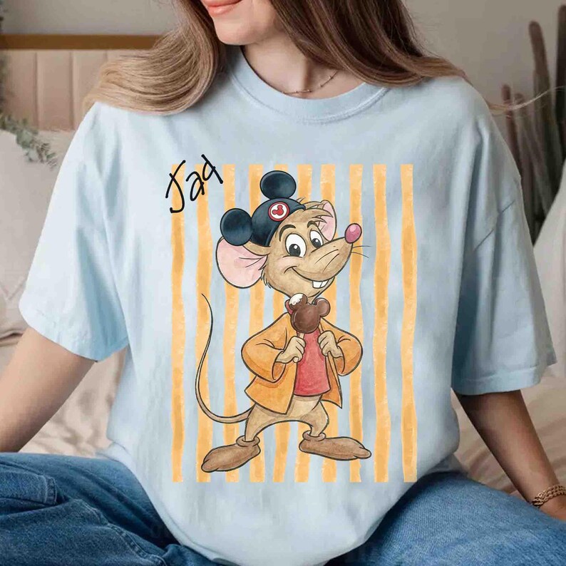 May include: A light blue t-shirt featuring a graphic of a cartoon mouse wearing a black hat with ears, a yellow jacket, and a red shirt, holding a brown treat. The text "Jad" is printed in black script above the mouse, set against yellow stripes.