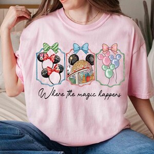 May include: Pink t-shirt featuring three framed designs with bows and Mickey Mouse ears, a globe, and balloons. The text "Where the magic happens" is written in cursive below the designs.