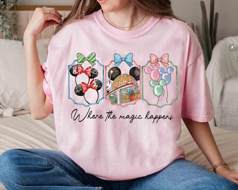 May include: Pink t-shirt featuring three framed designs with bows and Mickey Mouse ears, a globe, and balloons. The text "Where the magic happens" is written in cursive below the designs.