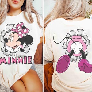 May include: Cream-colored t-shirt featuring a Minnie Mouse graphic. The front design shows Minnie bursting through a torn-paper effect, with the name "MINNIE" below. The back of the shirt shows Minnie's feet and a portion of her body.