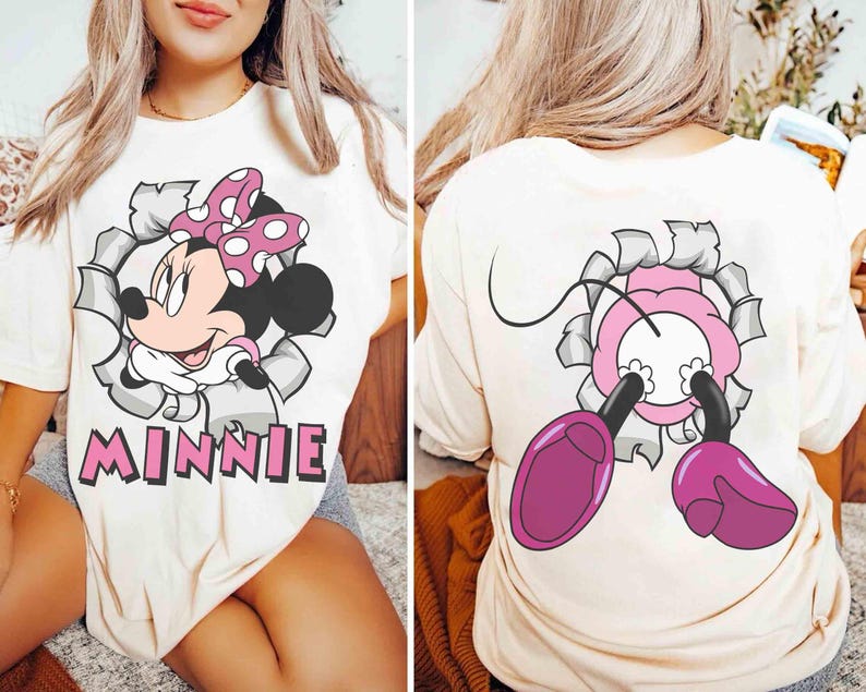 May include: Cream-colored t-shirt featuring a Minnie Mouse graphic. The front design shows Minnie bursting through a torn-paper effect, with the name "MINNIE" below. The back of the shirt shows Minnie's feet and a portion of her body.