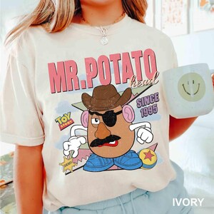 May include: Ivory-colored t-shirt featuring a graphic of Mr. Potato Head from Toy Story, wearing a cowboy hat and eye patch. The shirt includes the text "MR. POTATO head" and "SINCE 1995". A white mug with a smiley face is also visible.