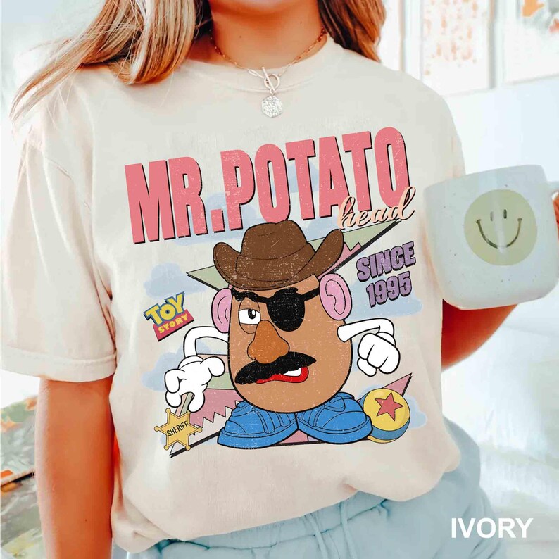 May include: Ivory-colored t-shirt featuring a graphic of Mr. Potato Head from Toy Story, wearing a cowboy hat and eye patch. The shirt includes the text "MR. POTATO head" and "SINCE 1995". A white mug with a smiley face is also visible.