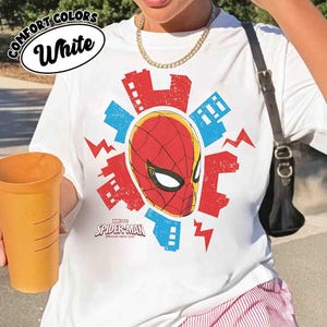 Puede incluir: Una camiseta blanca con un gr&aacute;fico central de la m&aacute;scara de Spider-Man en rojo y negro, rodeada de siluetas de edificios urbanos en rojo y azul y rayos. El texto Spider-Man Brand New Day est&aacute; impreso debajo de la m&aacute;scara.