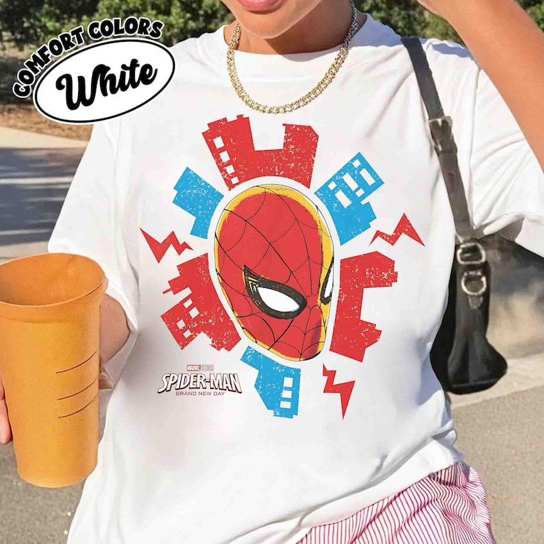 Puede incluir: Una camiseta blanca con un gr&aacute;fico central de la m&aacute;scara de Spider-Man en rojo y negro, rodeada de siluetas de edificios urbanos en rojo y azul y rayos. El texto Spider-Man Brand New Day est&aacute; impreso debajo de la m&aacute;scara.