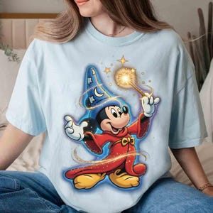 May include: Light blue t-shirt featuring a graphic of Mickey Mouse dressed as a wizard, holding a glowing wand. Mickey is wearing a red robe and a blue wizard hat with stars and moons. The design has a cartoon style.