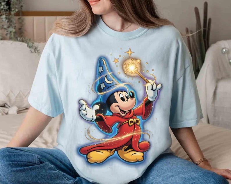 May include: Light blue t-shirt featuring a graphic of Mickey Mouse dressed as a wizard, holding a glowing wand. Mickey is wearing a red robe and a blue wizard hat with stars and moons. The design has a cartoon style.