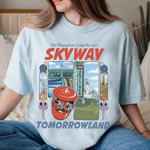 Puede incluir: Camiseta azul claro con un dise&ntilde;o gr&aacute;fico colorido del paseo Skyway de Disneyland con el texto "See Disneyland from the air! SKYWAY TOMORROWLAND". El dise&ntilde;o incluye im&aacute;genes del paseo y personajes.