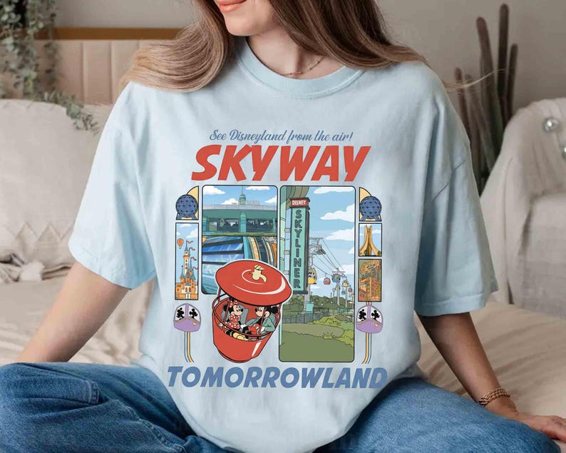 Puede incluir: Camiseta azul claro con un dise&ntilde;o gr&aacute;fico colorido del paseo Skyway de Disneyland con el texto "See Disneyland from the air! SKYWAY TOMORROWLAND". El dise&ntilde;o incluye im&aacute;genes del paseo y personajes.