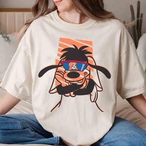 May include: A white t-shirt with a cartoon dog wearing sunglasses. The dog is wearing a red and white striped shirt and has a brown nose and black eyes. The dog is surrounded by orange and white stripes.
