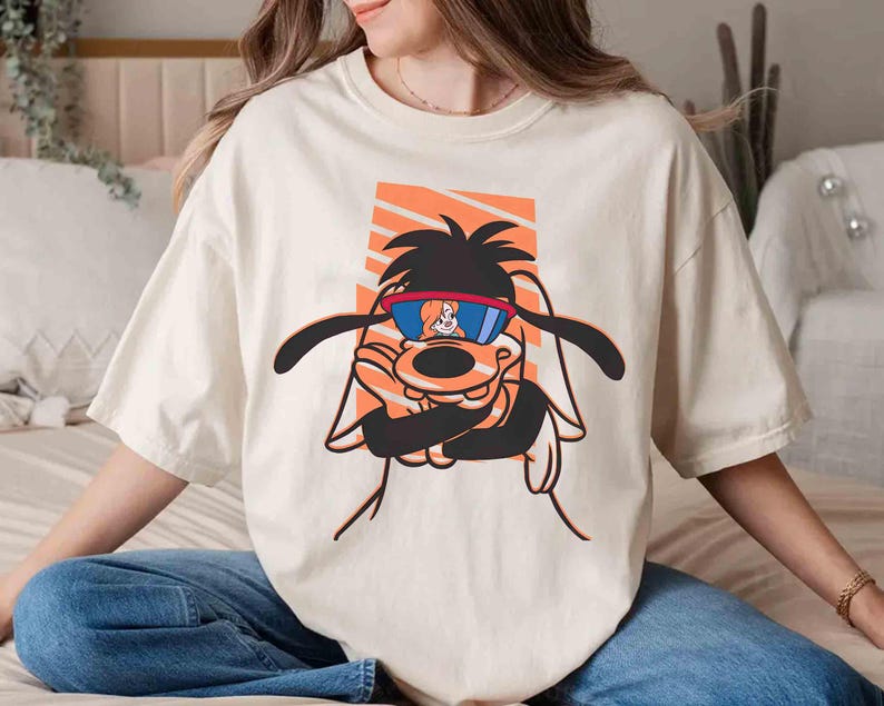May include: A white t-shirt with a cartoon dog wearing sunglasses. The dog is wearing a red and white striped shirt and has a brown nose and black eyes. The dog is surrounded by orange and white stripes.