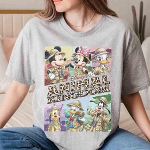 K&ouml;nnte beinhalten: Ein graues T-Shirt mit einem farbenfrohen Aufdruck der Disney-Figuren Mickey Mouse, Minnie Mouse, Donald Duck, Daisy Duck, Goofy und Pluto in Safari-Outfits. Der Text "Disney's Animal Kingdom" ist ebenfalls abgebildet.