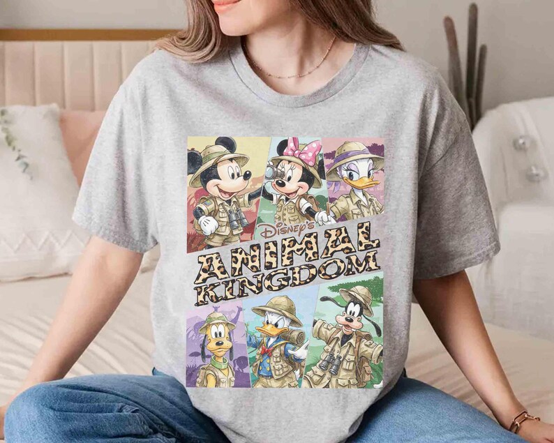 K&ouml;nnte beinhalten: Ein graues T-Shirt mit einem farbenfrohen Aufdruck der Disney-Figuren Mickey Mouse, Minnie Mouse, Donald Duck, Daisy Duck, Goofy und Pluto in Safari-Outfits. Der Text "Disney's Animal Kingdom" ist ebenfalls abgebildet.