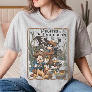 May include: A gray t-shirt featuring a graphic from Disney's "Pirates of the Caribbean." The design includes Mickey Mouse, Donald Duck, and other characters in pirate attire, with a treasure chest and a pirate flag. The text "Pirates of the Caribbean" is also visible.