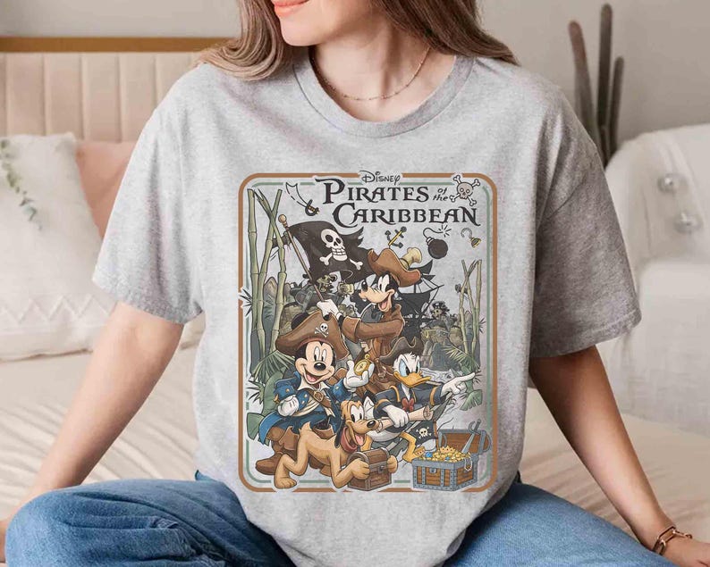 May include: A gray t-shirt featuring a graphic from Disney's "Pirates of the Caribbean." The design includes Mickey Mouse, Donald Duck, and other characters in pirate attire, with a treasure chest and a pirate flag. The text "Pirates of the Caribbean" is also visible.