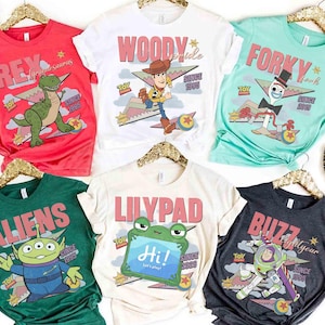 May include: A collection of colorful t-shirts featuring characters from the Toy Story franchise. Designs include Rex, Woody, Forky, Aliens, Lilypad, and Buzz Lightyear, with text and graphics. Each shirt is displayed on a gold hanger.