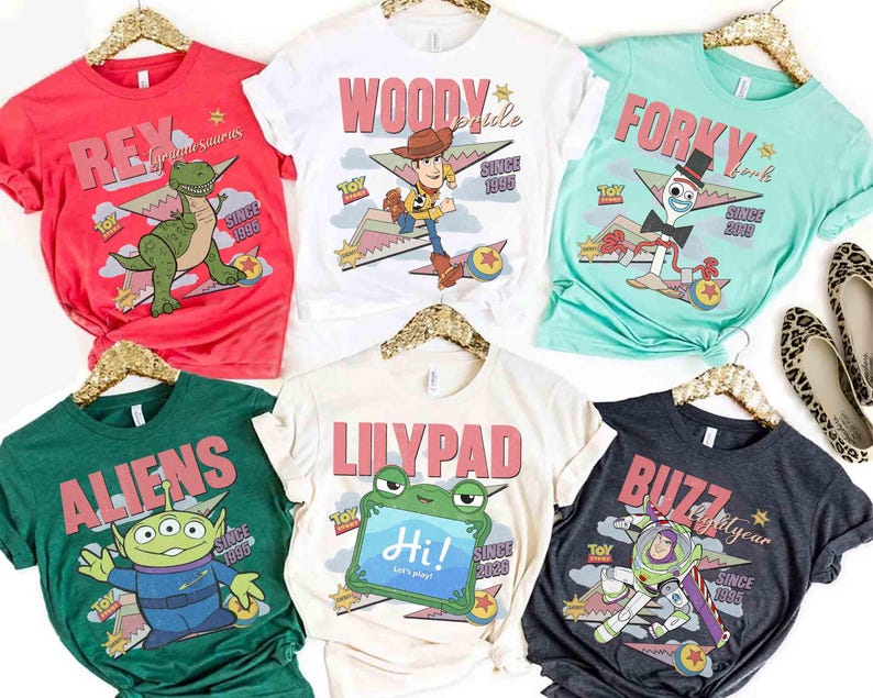 May include: A collection of colorful t-shirts featuring characters from the Toy Story franchise. Designs include Rex, Woody, Forky, Aliens, Lilypad, and Buzz Lightyear, with text and graphics. Each shirt is displayed on a gold hanger.