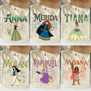Op de afbeelding: Zes cr&egrave;mekleurige t-shirts met verschillende Disney-prinsessenontwerpen. Elk shirt heeft de naam van een prinses in een decoratief lettertype boven een illustratie van de prinses. De prinsessen zijn Anna, Merida, Tiana, Mulan, Rapunzel en Moana.