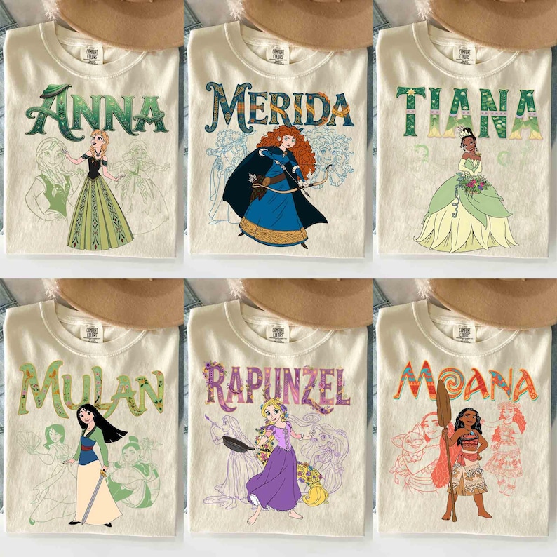 Op de afbeelding: Zes cr&egrave;mekleurige t-shirts met verschillende Disney-prinsessenontwerpen. Elk shirt heeft de naam van een prinses in een decoratief lettertype boven een illustratie van de prinses. De prinsessen zijn Anna, Merida, Tiana, Mulan, Rapunzel en Moana.
