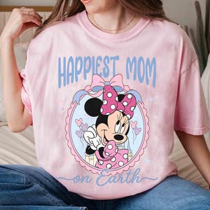 Pode incluir: Camiseta rosa claro com a frase "Happiest Mom on Earth" em azul claro. O design apresenta Minnie Mouse com um la&ccedil;o rosa e bolinhas, emoldurada em um oval.