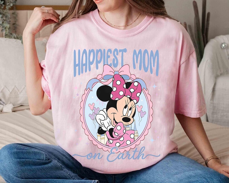 Pode incluir: Camiseta rosa claro com a frase "Happiest Mom on Earth" em azul claro. O design apresenta Minnie Mouse com um la&ccedil;o rosa e bolinhas, emoldurada em um oval.