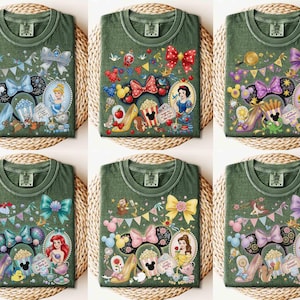 May include: Six olive green t-shirts with colorful Disney princess-themed designs. Each shirt features a different princess, such as Cinderella, Snow White, and Ariel, along with related imagery like bows, shoes, and teacups. The shirts are displayed on a woven surface.