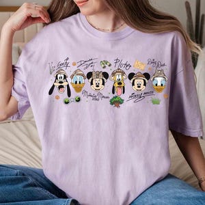May include: A light purple t-shirt featuring cartoon characters Goofy, Donald Duck, Minnie Mouse, Pluto, Mickey Mouse, and Daisy Duck. Each character is wearing a safari hat. The names of the characters are written above them.