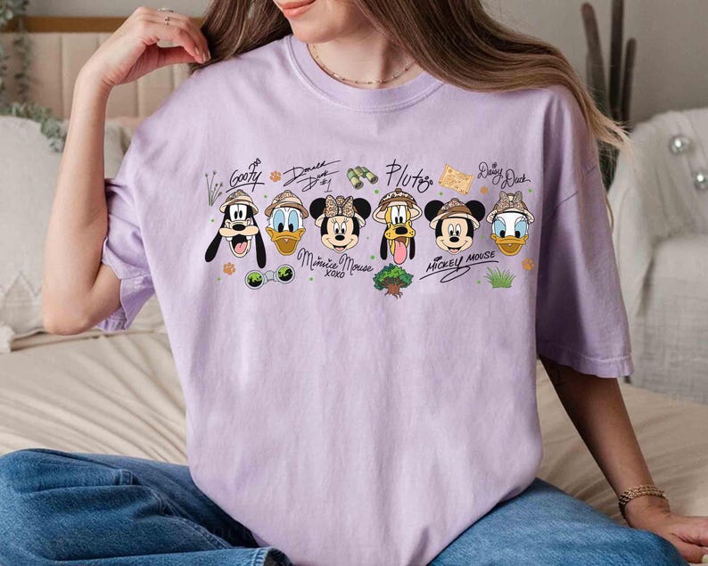 May include: A light purple t-shirt featuring cartoon characters Goofy, Donald Duck, Minnie Mouse, Pluto, Mickey Mouse, and Daisy Duck. Each character is wearing a safari hat. The names of the characters are written above them.