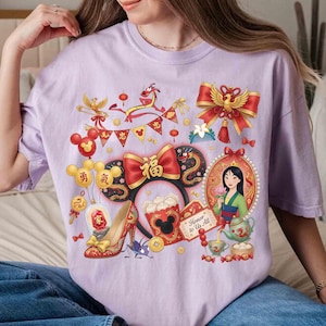 May include: A lilac-colored t-shirt featuring a colorful graphic design. The design includes a portrait of a woman, a red bow, a dragon, and other festive elements. The words "Honor to Us All" are also visible.
