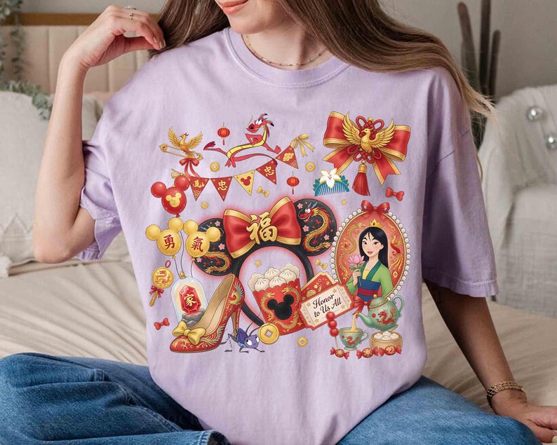 May include: A lilac-colored t-shirt featuring a colorful graphic design. The design includes a portrait of a woman, a red bow, a dragon, and other festive elements. The words "Honor to Us All" are also visible.
