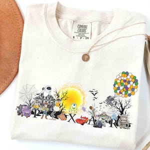 May include: Cream-colored t-shirt featuring a colorful Pixar-themed graphic. The design includes various characters, a yellow sun, and the word "PIXAR" in large letters. A gold necklace with a pendant is draped on the shirt.