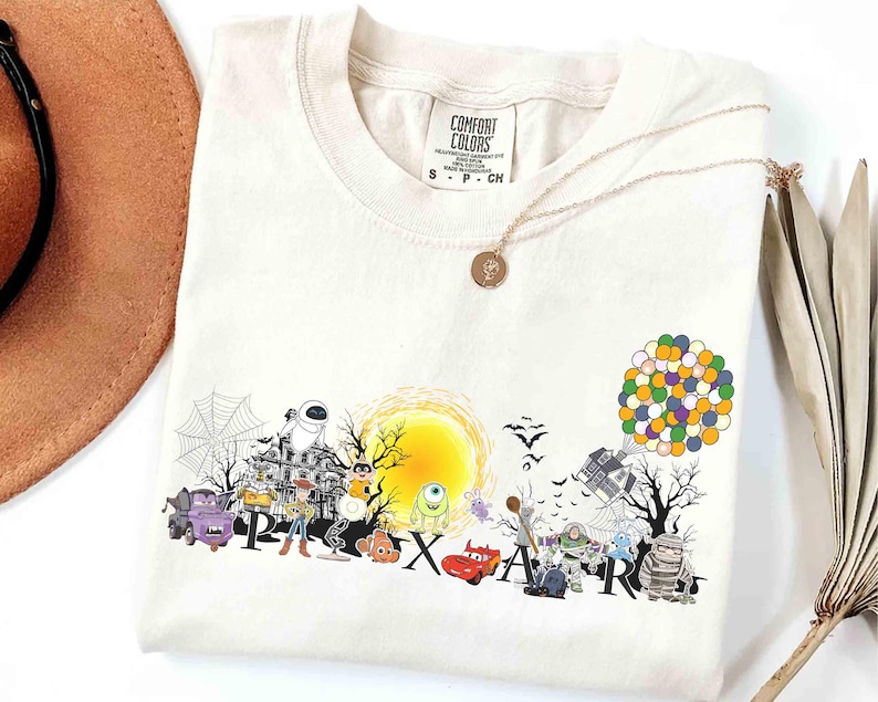 May include: Cream-colored t-shirt featuring a colorful Pixar-themed graphic. The design includes various characters, a yellow sun, and the word "PIXAR" in large letters. A gold necklace with a pendant is draped on the shirt.