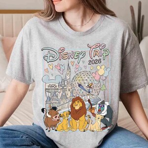 May include: A light gray t-shirt featuring a colorful graphic of Disney characters and landmarks. The design includes the text "Disney Trip 2026" and images of the Lion King characters, a castle, and balloons. The shirt is a crew neck.