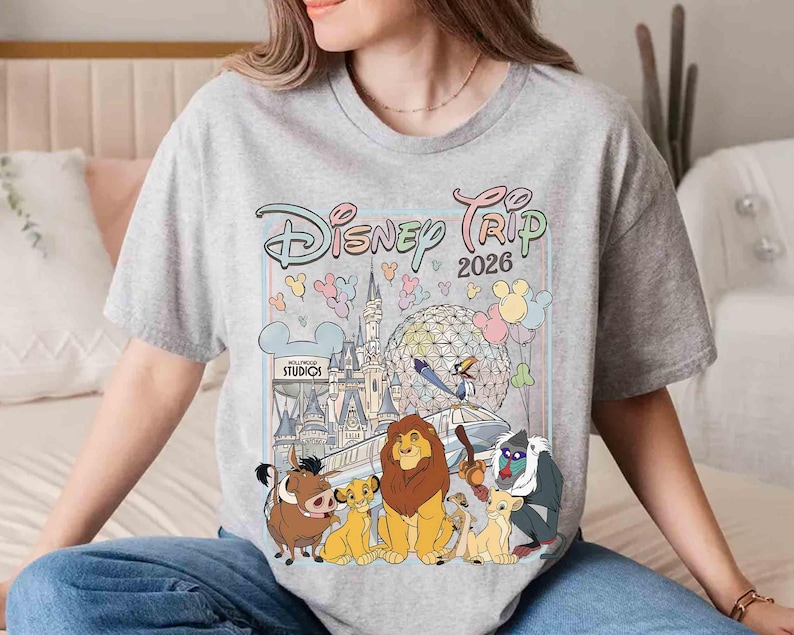 May include: A light gray t-shirt featuring a colorful graphic of Disney characters and landmarks. The design includes the text "Disney Trip 2026" and images of the Lion King characters, a castle, and balloons. The shirt is a crew neck.