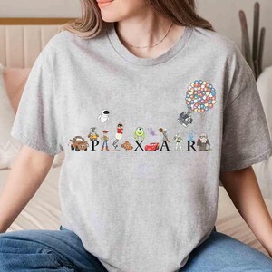 K&ouml;nnte beinhalten: Ein graues T-Shirt mit einem farbenfrohen Pixar-Design. Das Design zeigt verschiedene Charaktere, darunter Lightning McQueen und Wall-E, und das Wort "PIXAR". Eine Ansammlung von Ballons schwebt &uuml;ber dem "R" im Design.